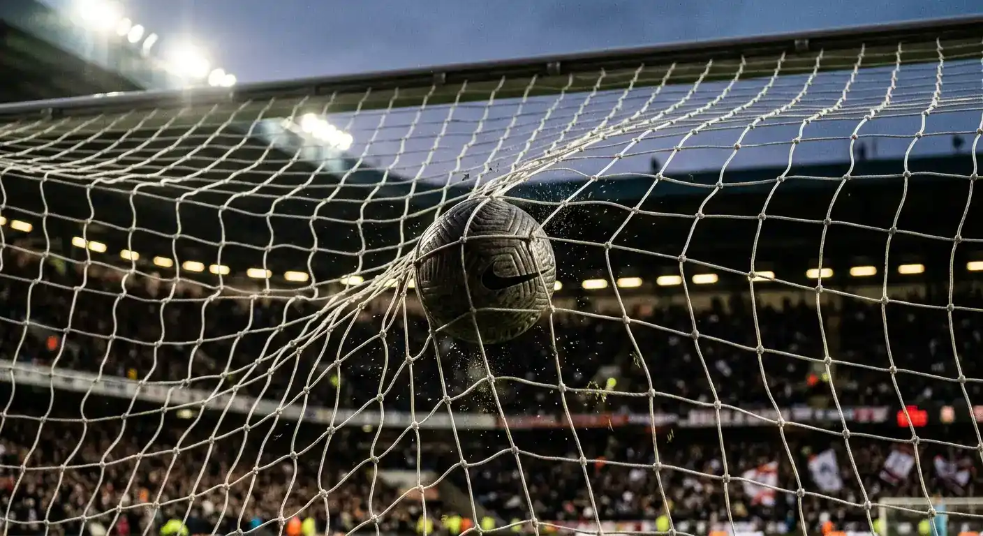 Nahaufnahme eines Fußballs im Netz eines Tores bei Flutlicht, im Hintergrund unscharf die Tribüne
